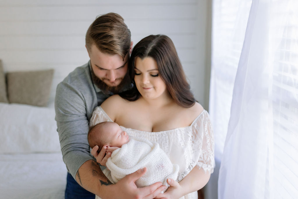 A mother cradles her sleeping newborn baby while standing by a window with dad hugging her from behind lavender lactation