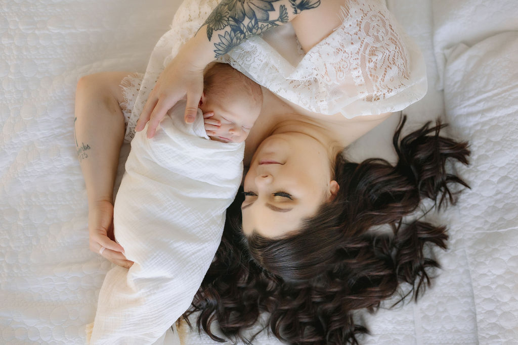 A mother lays with and cradles her sleeping newborn baby on a bed in a white lace dress