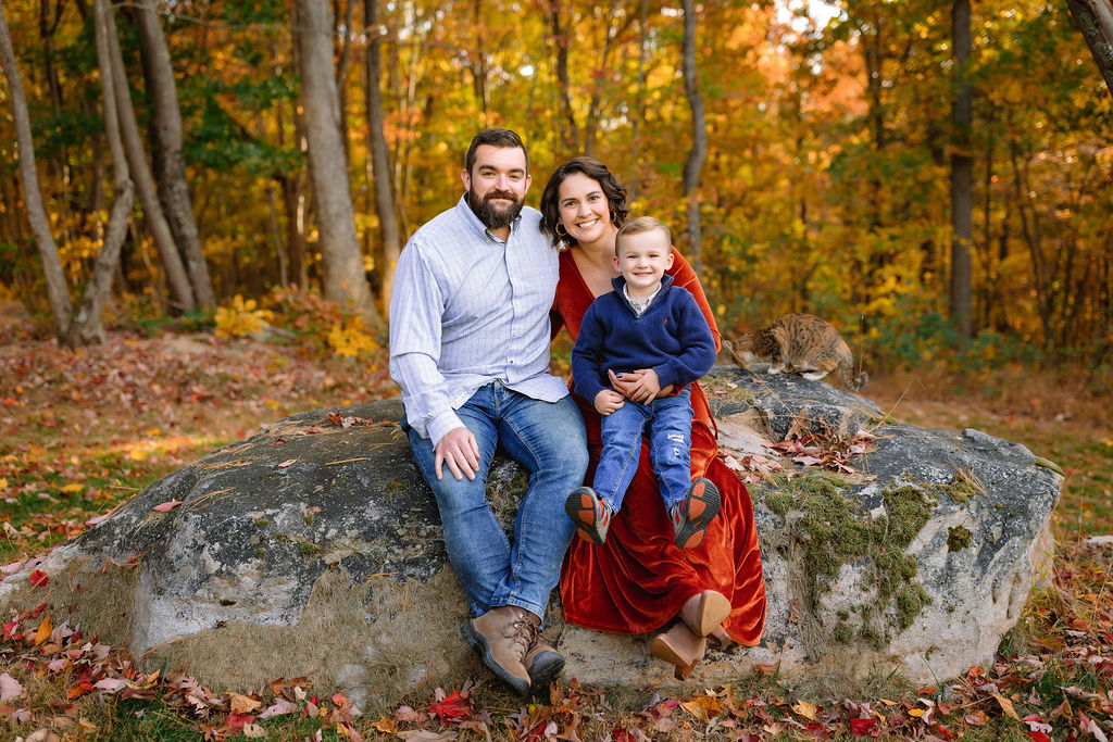 A family of three sit together on a large boulder in the woods