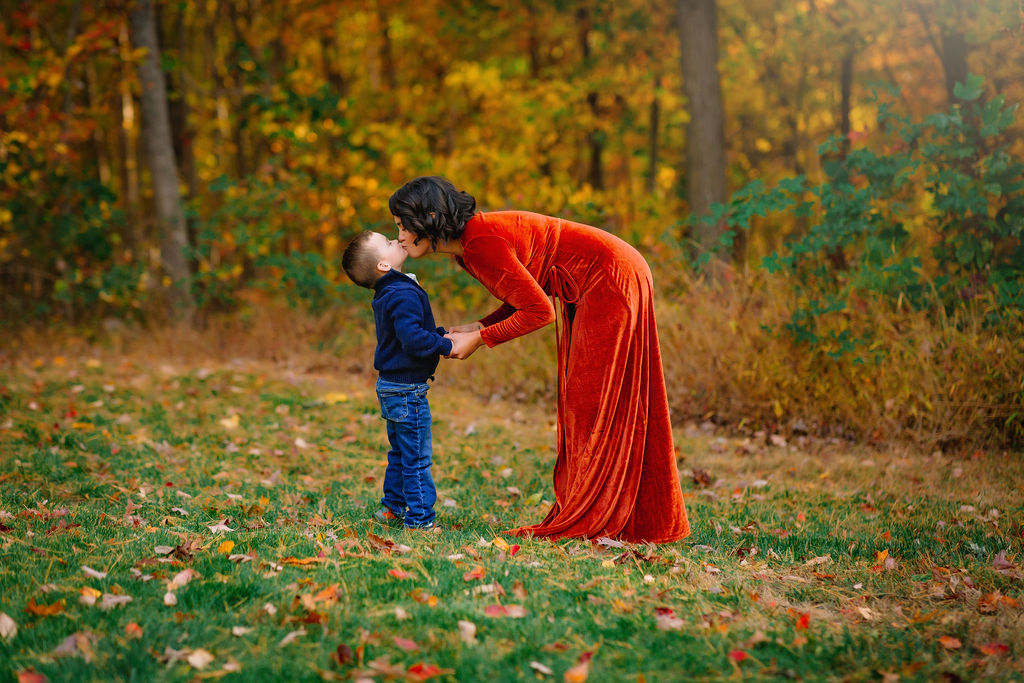 A mom in a red dress bends over to kiss her young son on the edge of a forest in fall montessori preschool charlottesville