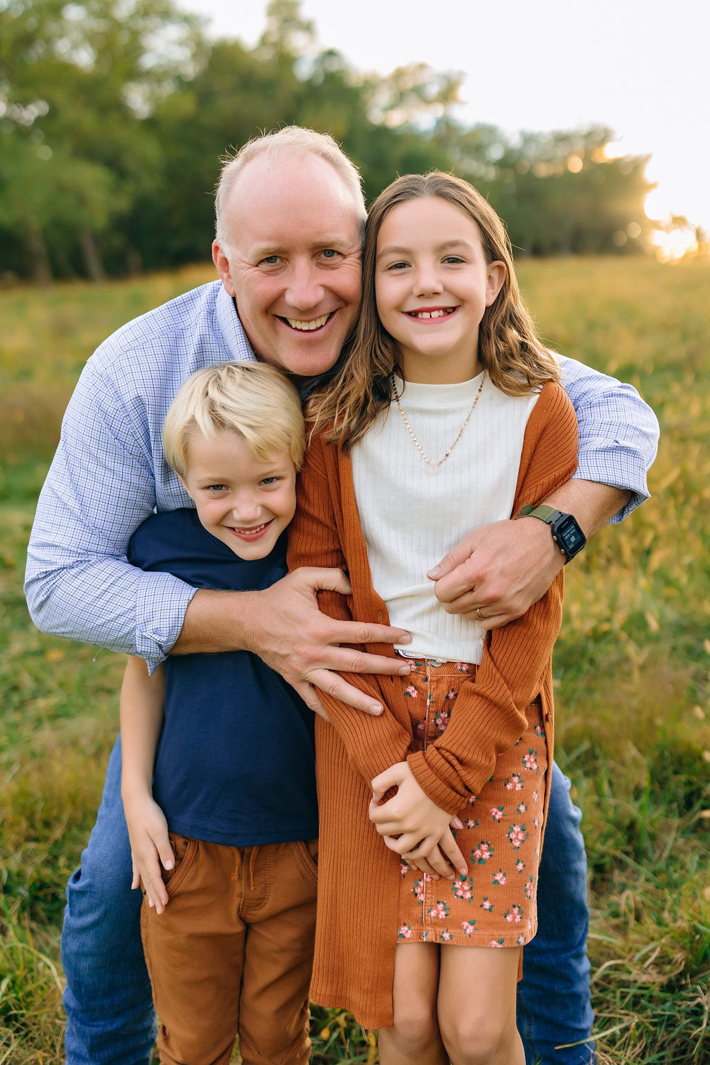 A father hugs onto his young son and daughter on a grassy hill at sunset