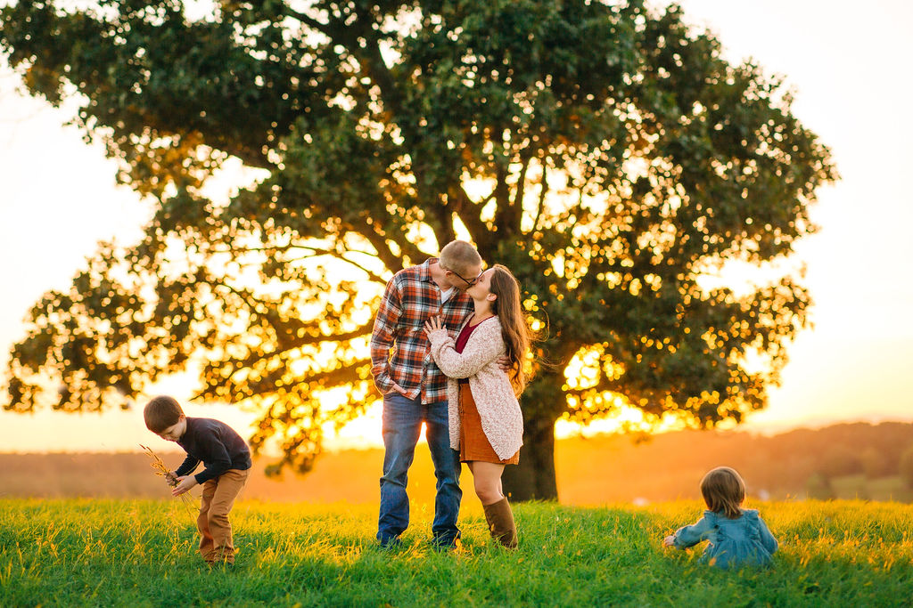 Mom and dad kiss while their two young children pick the grass around them in a pasture