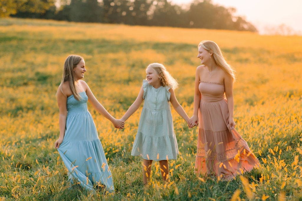 Three Sisters hold hands in dresses while walking through a field at sunset charlottesville apple picking
