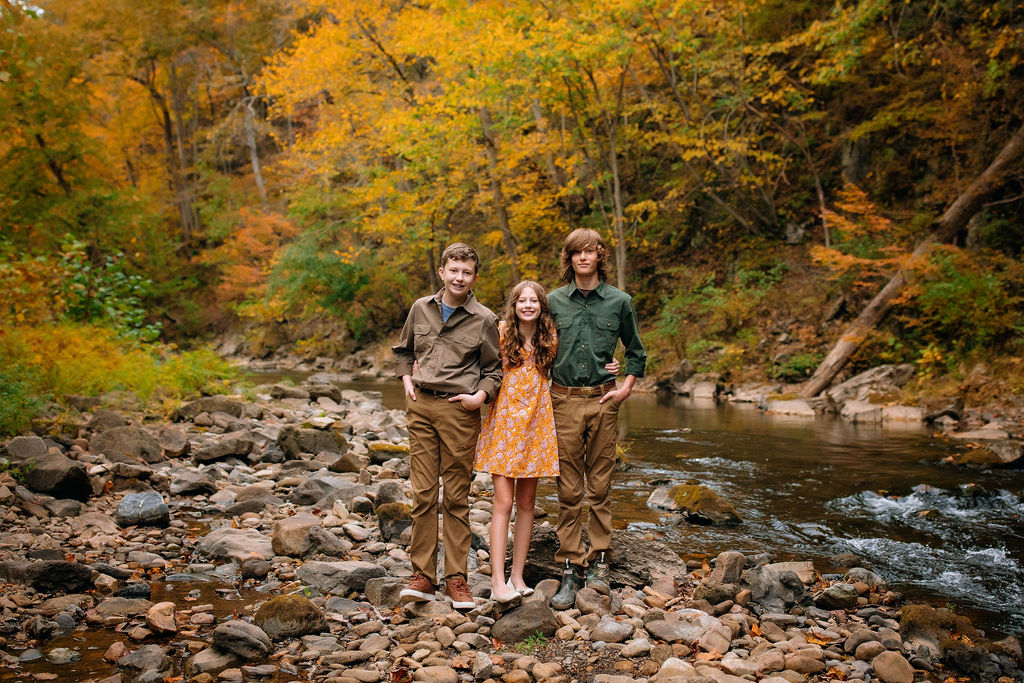 Three siblings stand on a river's edge with hands in pockets in fall