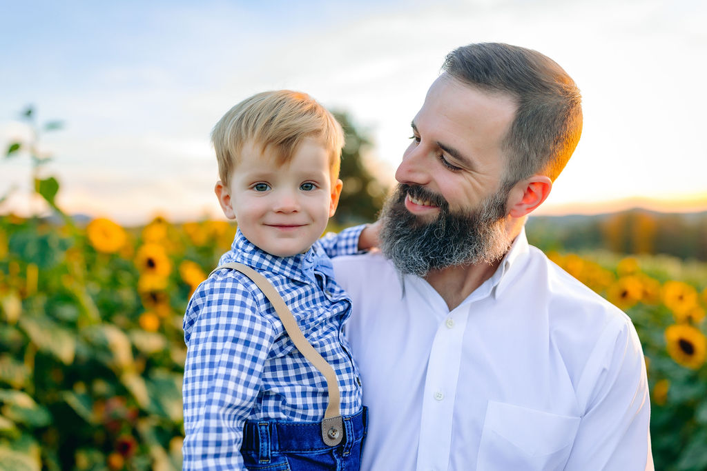 A father smiles at his young toddler son wearing blue shirt and jeans while standing in a sunflower field charlottesville pumpkin patch