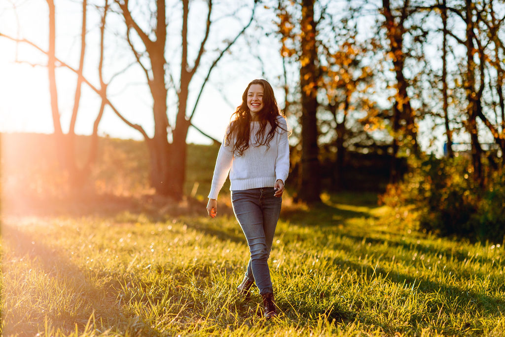 A teenage girl in a white sweater and jeans walks through a field at sunset harrisonburg fall festival
