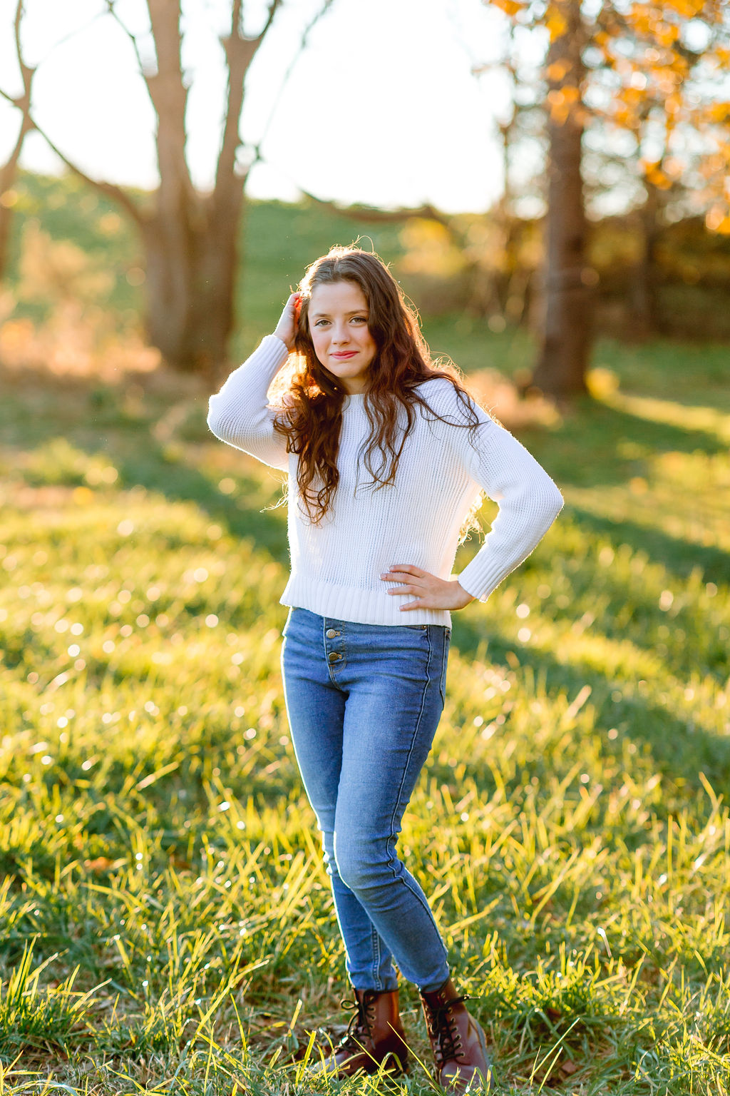 A teenage girl in jeans and a white sweater runs a hand through her hair in a field at sunset