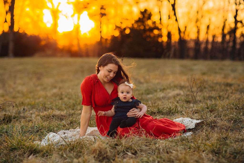 A young mom sits on a picnic blanket in a field at sunset with her infant daughter in her lap pumpkin patch near harrisonburg va