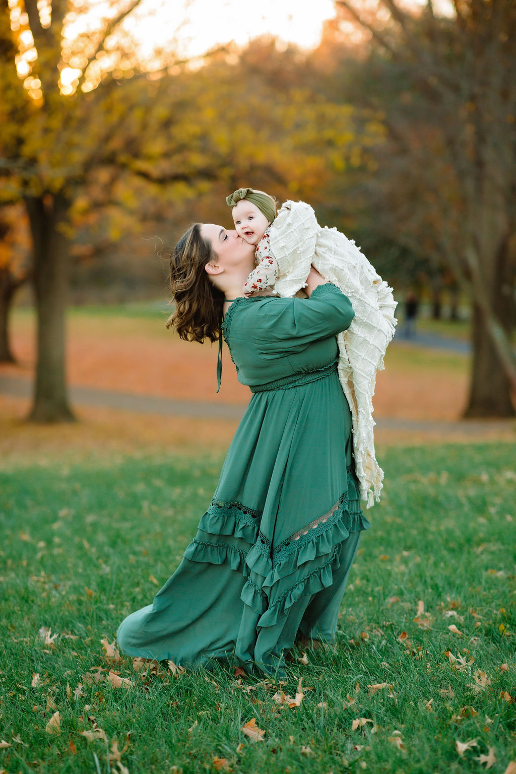 A new mom lifts and plays with her infant daughter while wearing a long green dress in a field at sunset