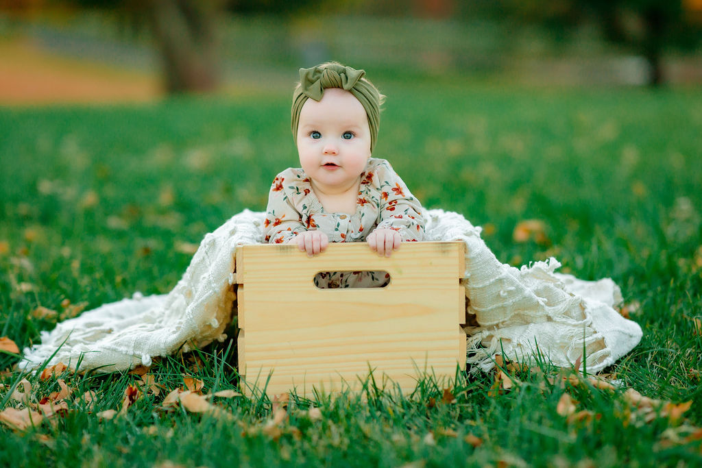 An infant girl in a floral dress sits in a wooden box in a grassy field waiting for Charlottesville christmas lights