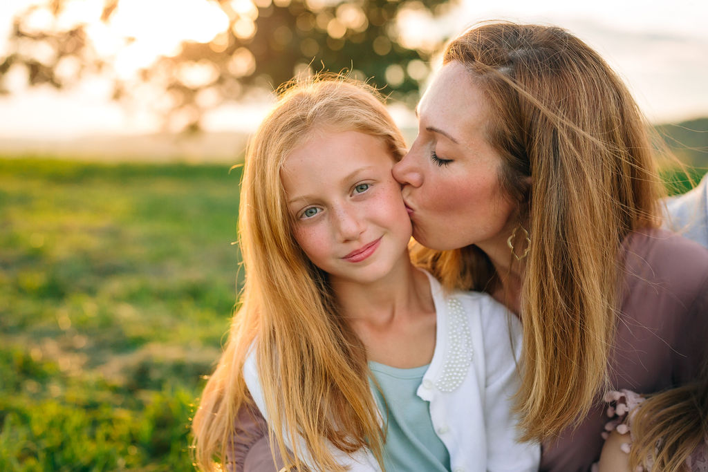 A mother in a pink dress kisses the cheek of her young daughter in a white sweater in a field at sunset before they visit charlottesville christmas events