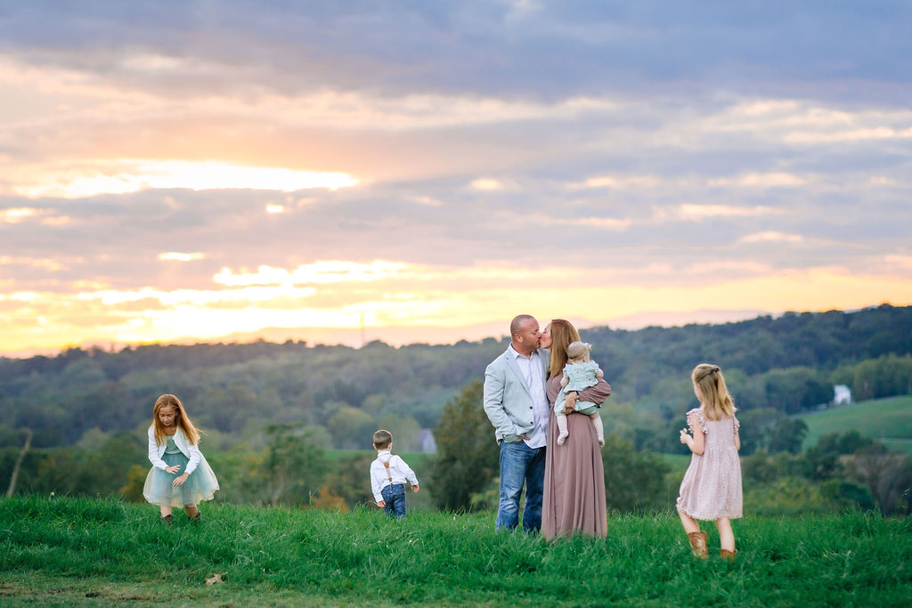 A mom holds her infant daughter on her hip while kissing her husband in a sports coat as they stand on a hill top with their three young kids playing around them at sunset before enjoying charlottesville christmas events