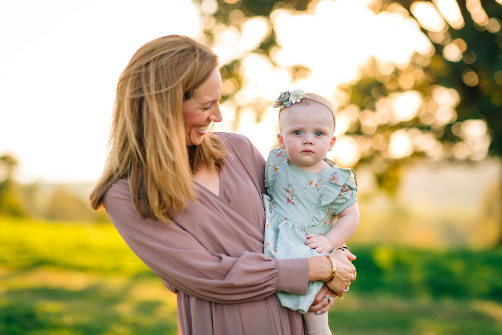 A mother in a purple dress holds her infant daughter in a green floral dress in a field at sunset