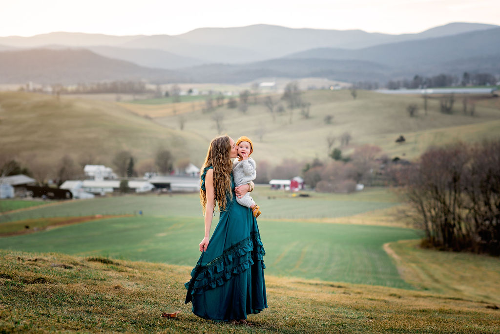 A mother in a blue dress kisses the cheek of her infant child while they walk through a field in the mountains