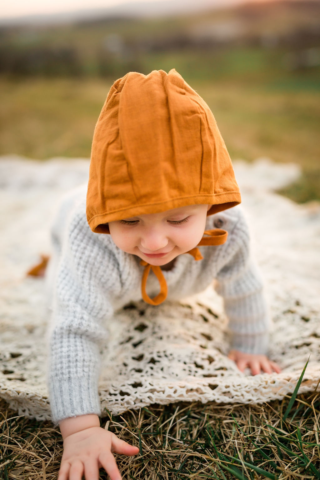 An infant crawls on a picnic blanket in a field in fall in an orange hat and grey sweater before visiting christmas tree farms in charlottesville va