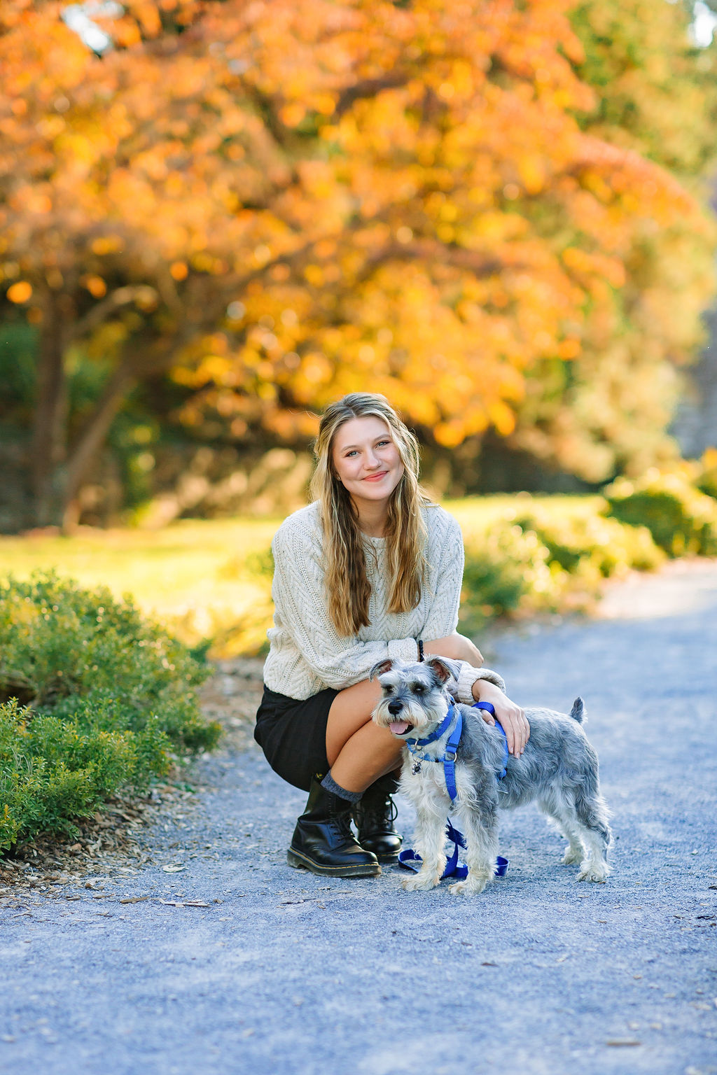 A teenage girl in a white sweater and black boots squats on a park path with her small grey puppy at sunset
