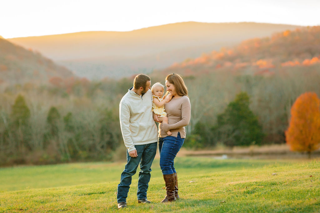 A mother in jeans and a tan shirt holds her infant daughter while standing in a field at sunset in fall with her husband at her side before visiting harrisonburg winter wonderfest
