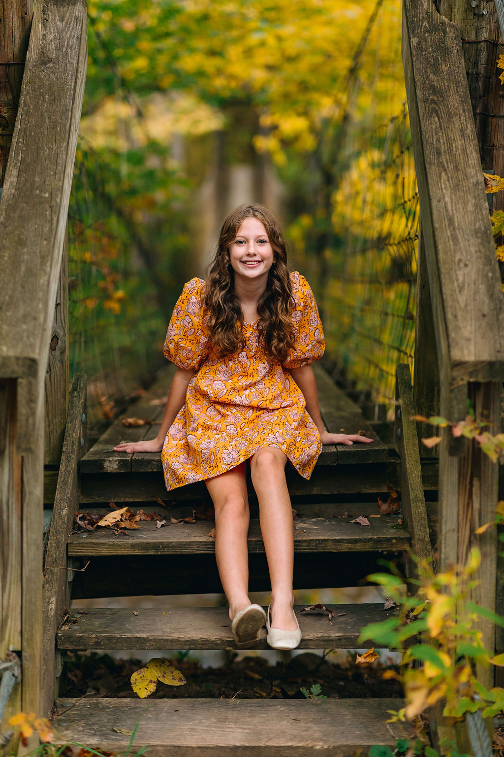 A young girl sits on the steps of a narrow wooden bridge in the woods in fall in an orange floral dress