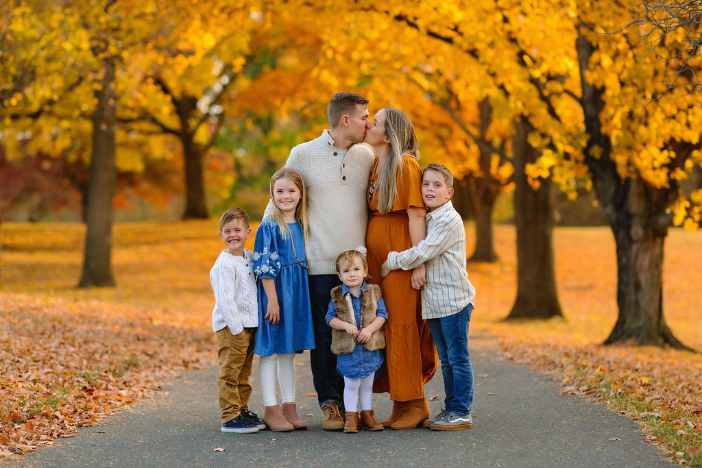 Mom and dad kiss while standing in a park path in full fall colors with their four children hugging them
