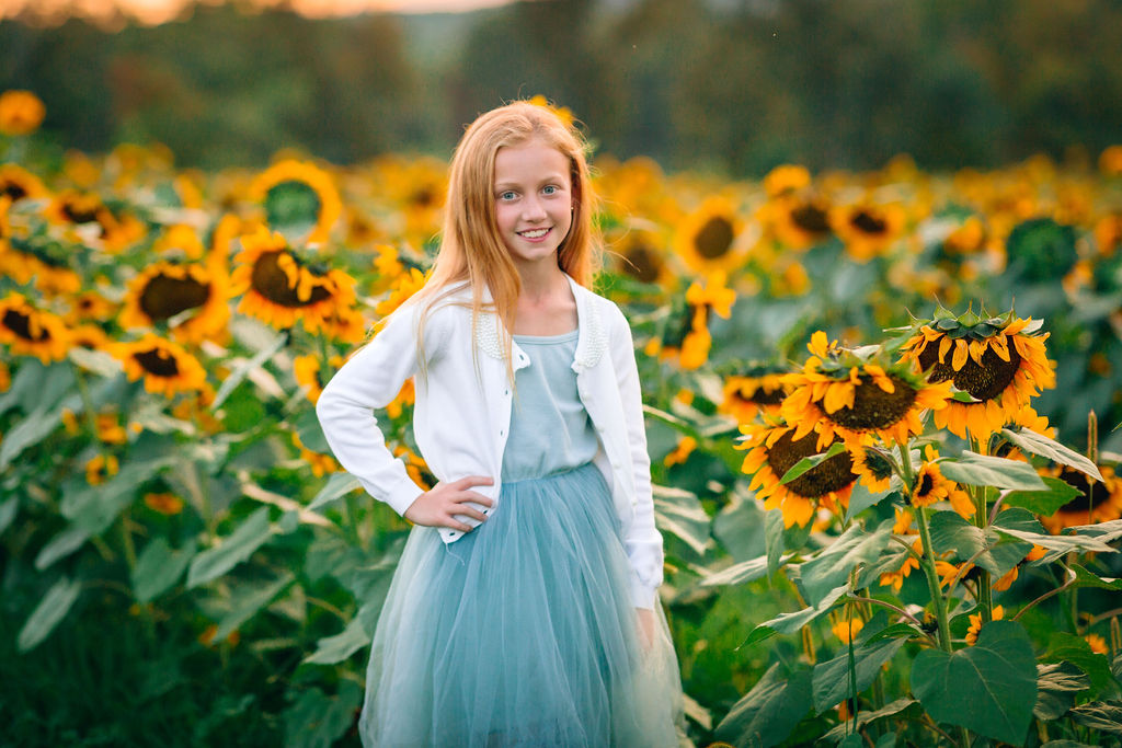 A young girl in a blue dress and white sweater stands in a field of sunflowers at a staunton va pumpkin patch
