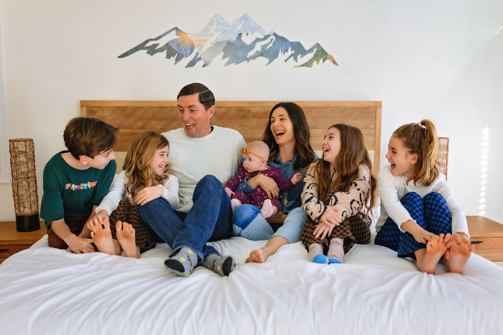 A family off seven laugh and sit together on a bed with the kids in pajamas