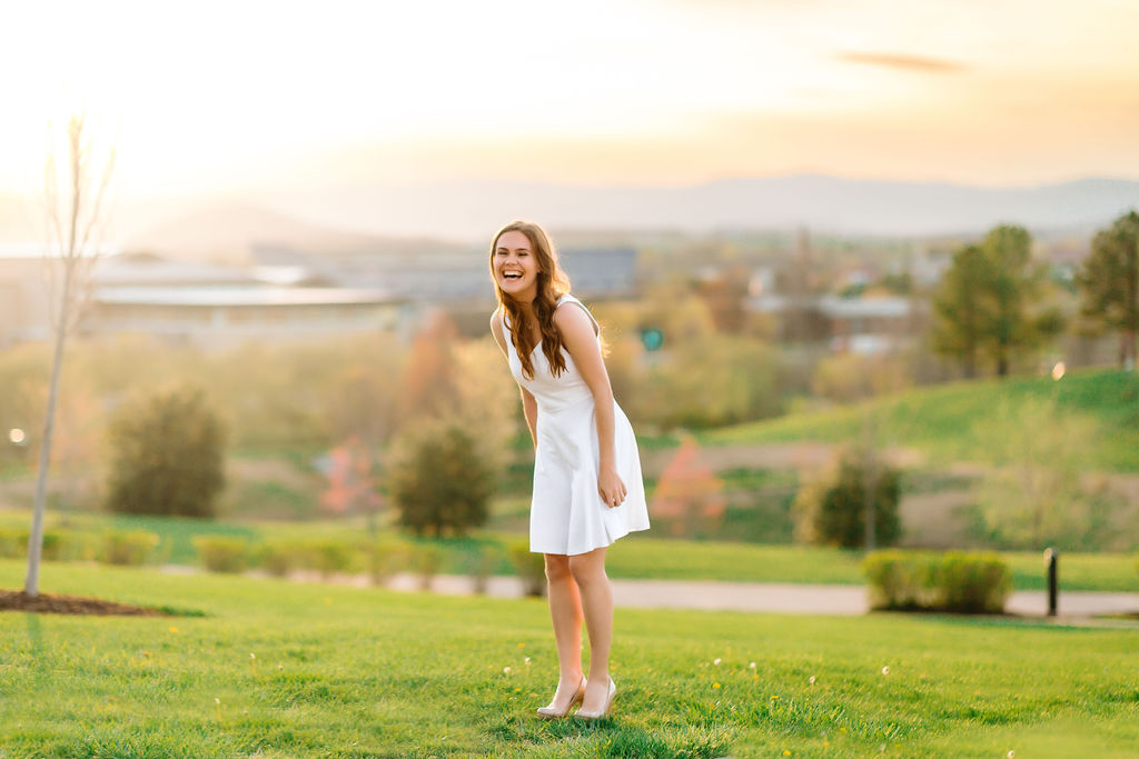 A college grad stands on a grassy hill in a white dress laughing after completing her jmu bucket list