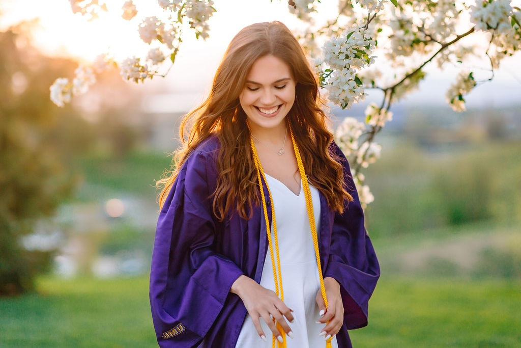 A college grad in a white dress and purple gown plays with her gold tassels under a flowering tree