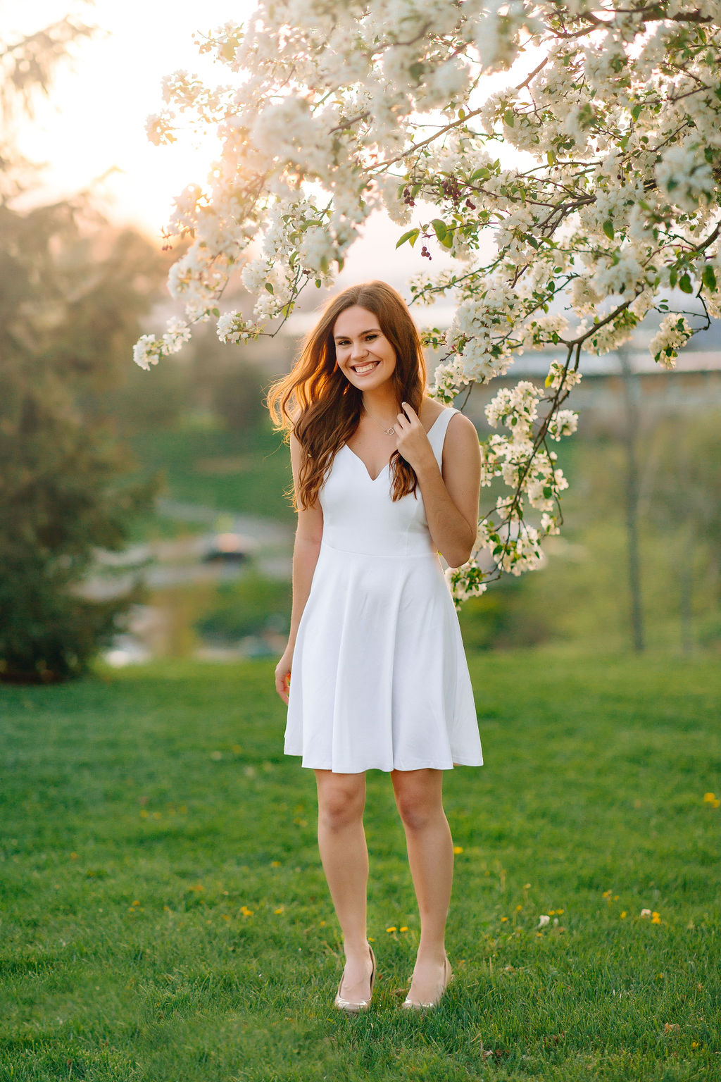 A college grad in a white dress stands under a flowering tree at sunset after taking jmu dual enrollment courses