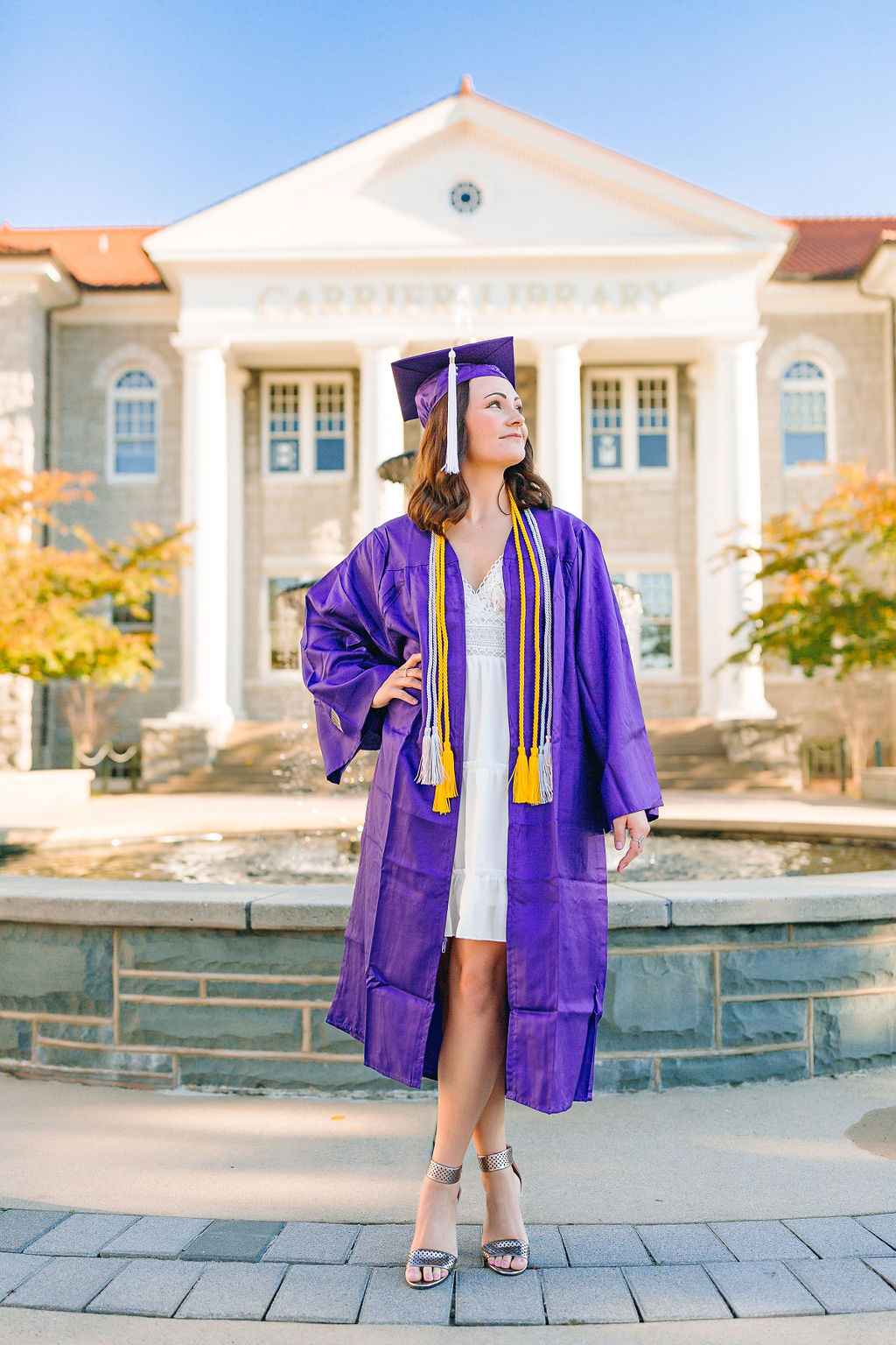 A college grad stands with a hand on her hip in front of a fountain in a purple cap and gown