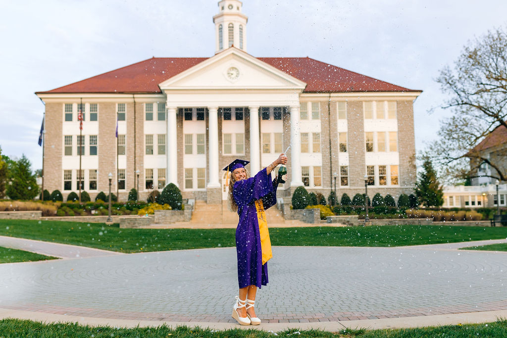 A college graduate pops a bottle of champagne on a brick patio in her purple gown before starting a jmu graduate program