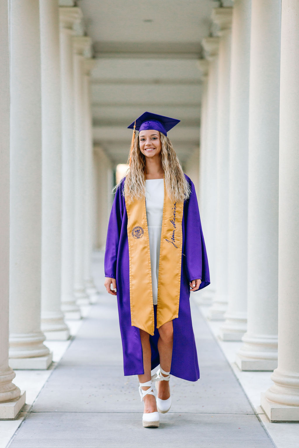 A college graduate walks through a covered path in a purple and gold cap and gown before starting a jmu graduate program