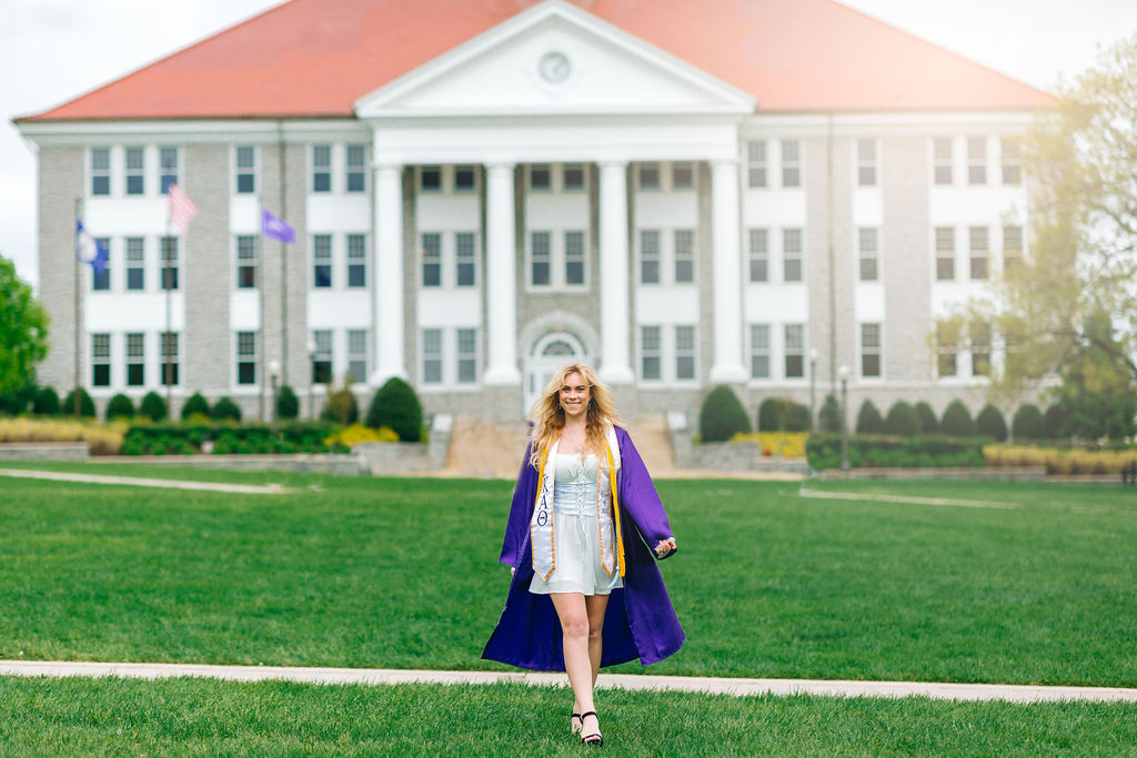 A college graduate in a blue dress walks through the grass during jmu graduation photos in her purple gown