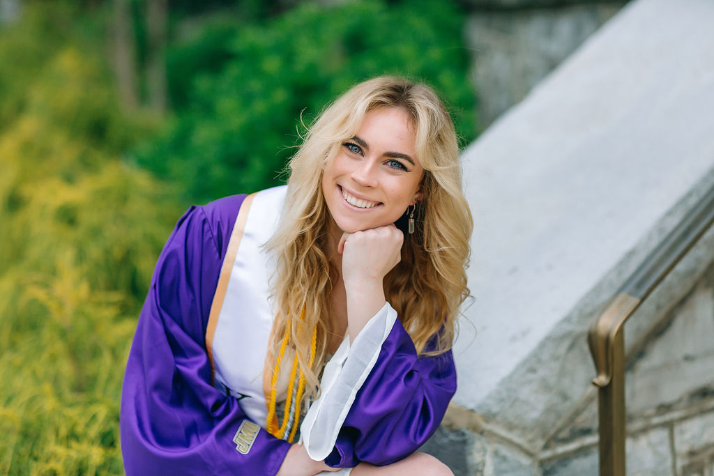A college graduate rests her head on her chin by a stone staircase in her purple gown