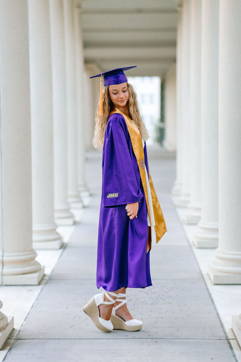 A graduate in her purple cap and gown stands in a row of columns looking down her shoulder for jmu graduation photos