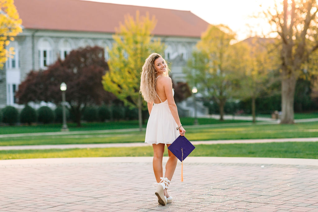 A college graduate walks through the quad holding her purple cap and wearing a white dress for jmu graduation photos