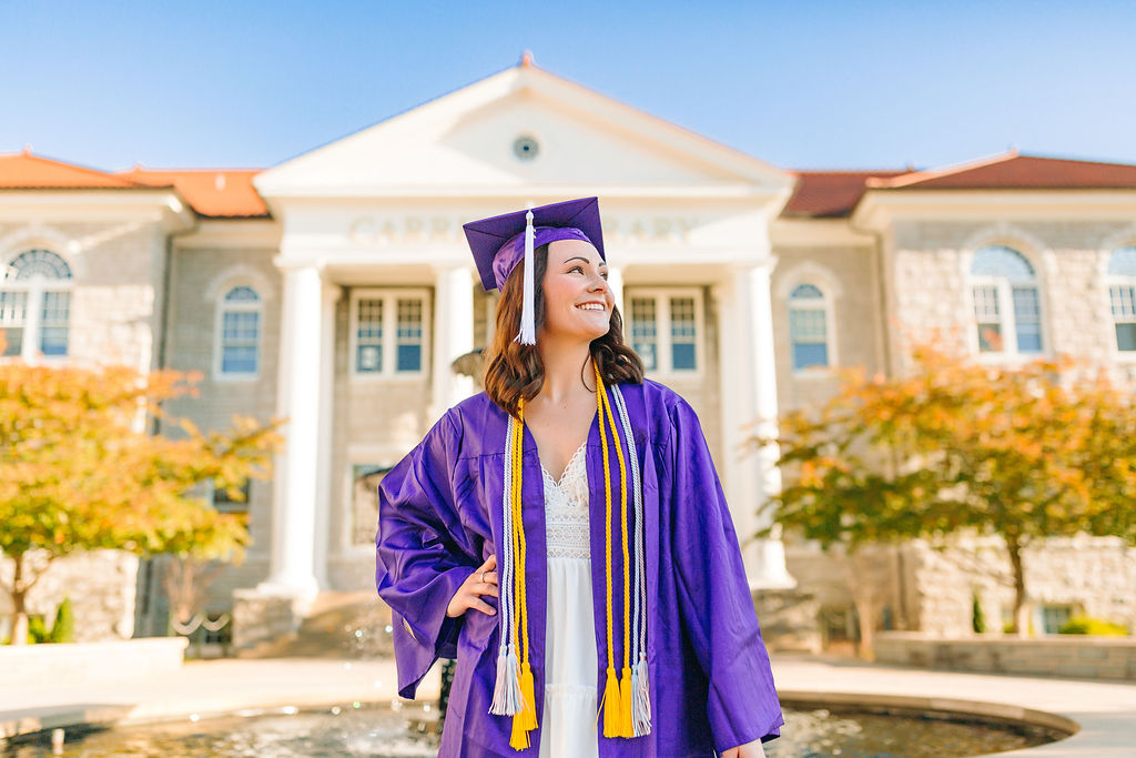 A college graduate stands smiling in front of jmu on campus housing in her purple cap and gown