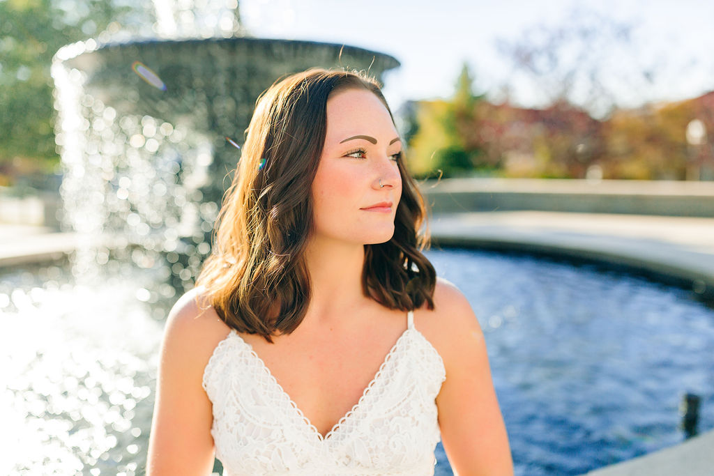 A college graduate stands in front of a fountain looking over her shoulder in a white dress