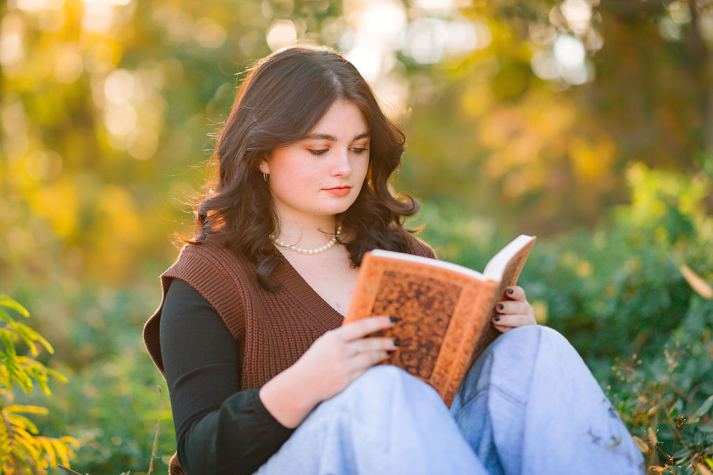 A high school senior sits in a forest at sunset in a brown sweater and jeans reading a book while waiting for jmu scholarships