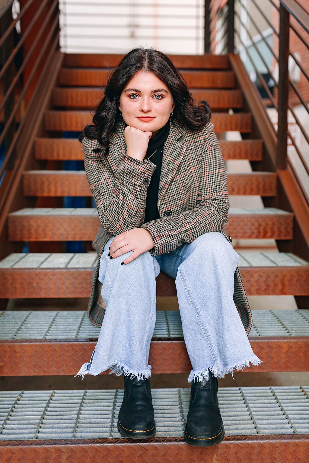 A high school senior sits in a plaid coat and jeans on some rusted stairs