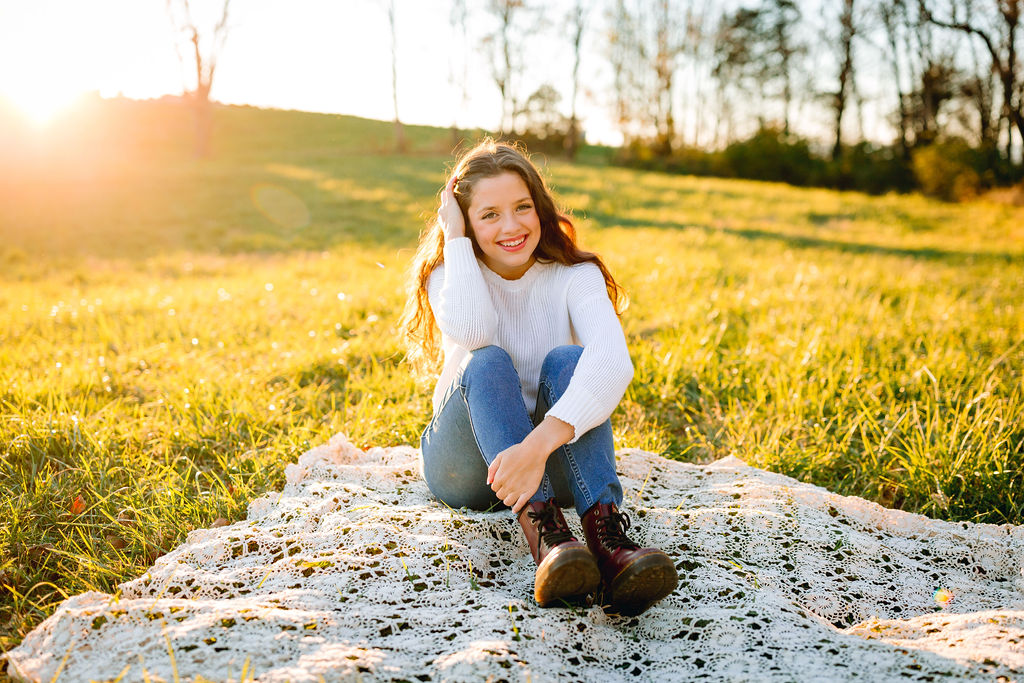A graduate sits on a lace blanket in a grassy field at sunset before joining jmu student organizations