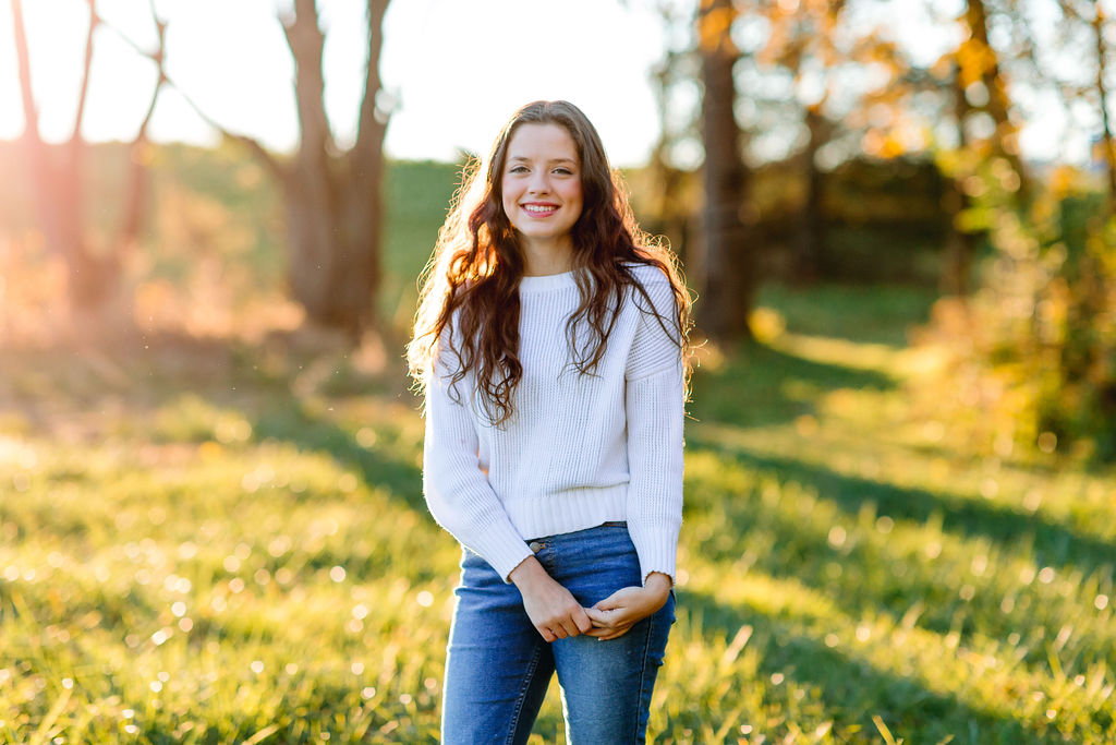 A high school graduate stands in a white sweater and jeans in a grassy field