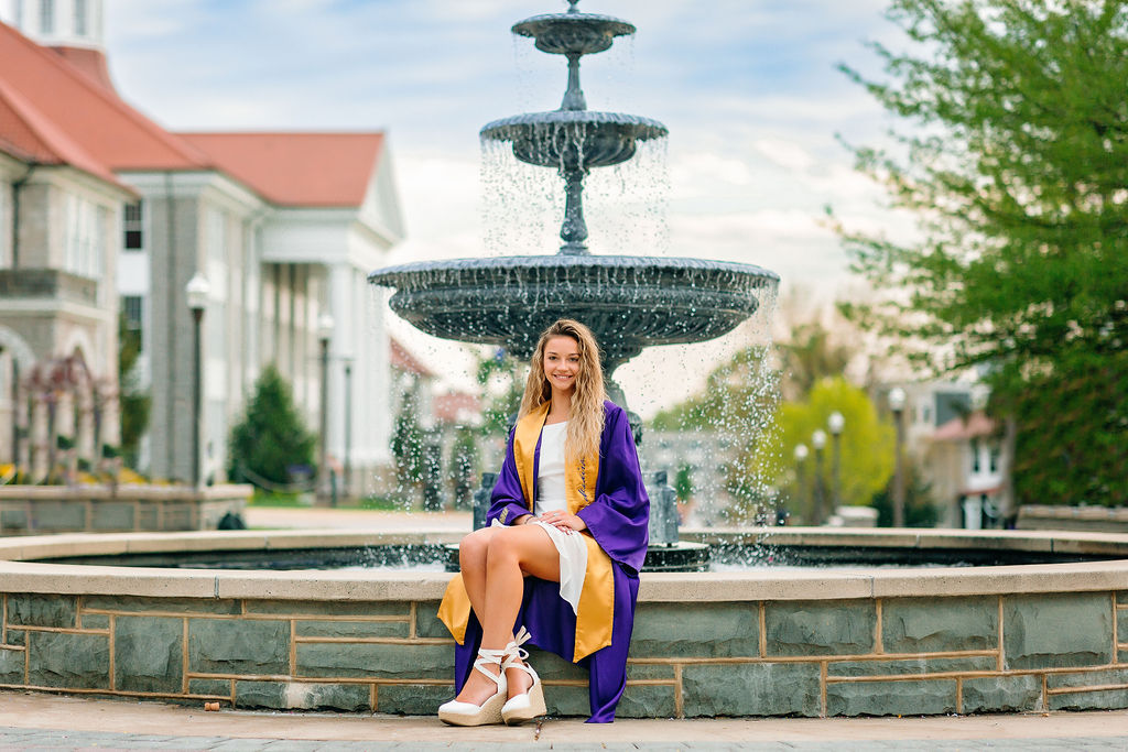 A college grad in a white dress and purple gown sits on the edge of a fountain