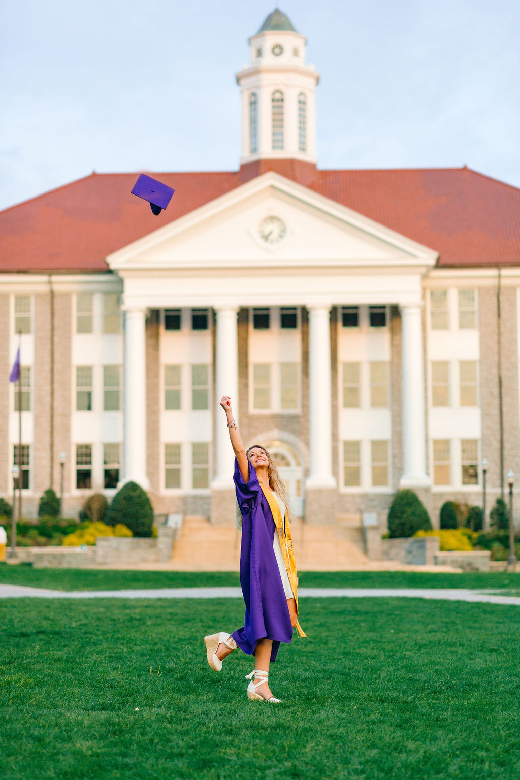 A college grad in a white dress and purple gown tosses her cap in the air while standing in a lawn thanks to jmu tutoring