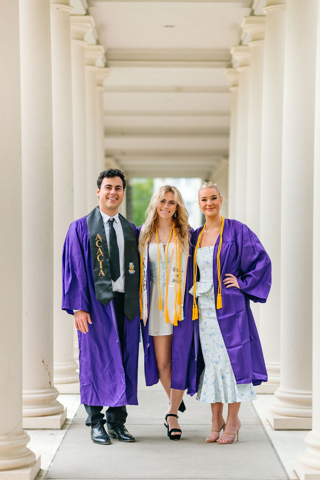 Three college graduates stand together in a row of columns wearing their purple robes in jmu writing center