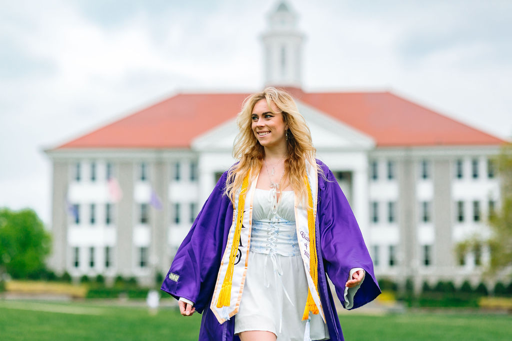 A college graduate walks through campus in a purple robe and blue dress while smiling at jmu writing center