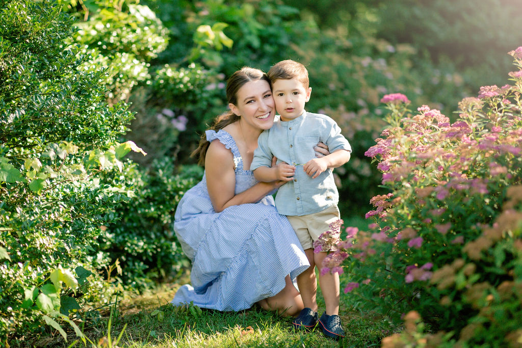 A mom kneels in a flower garden at sunset hugging her toddler son in a blue shirt