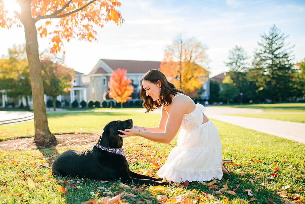 A girl in a white dress plays with her black dog in a park lawn under a tree while visiting JMU
